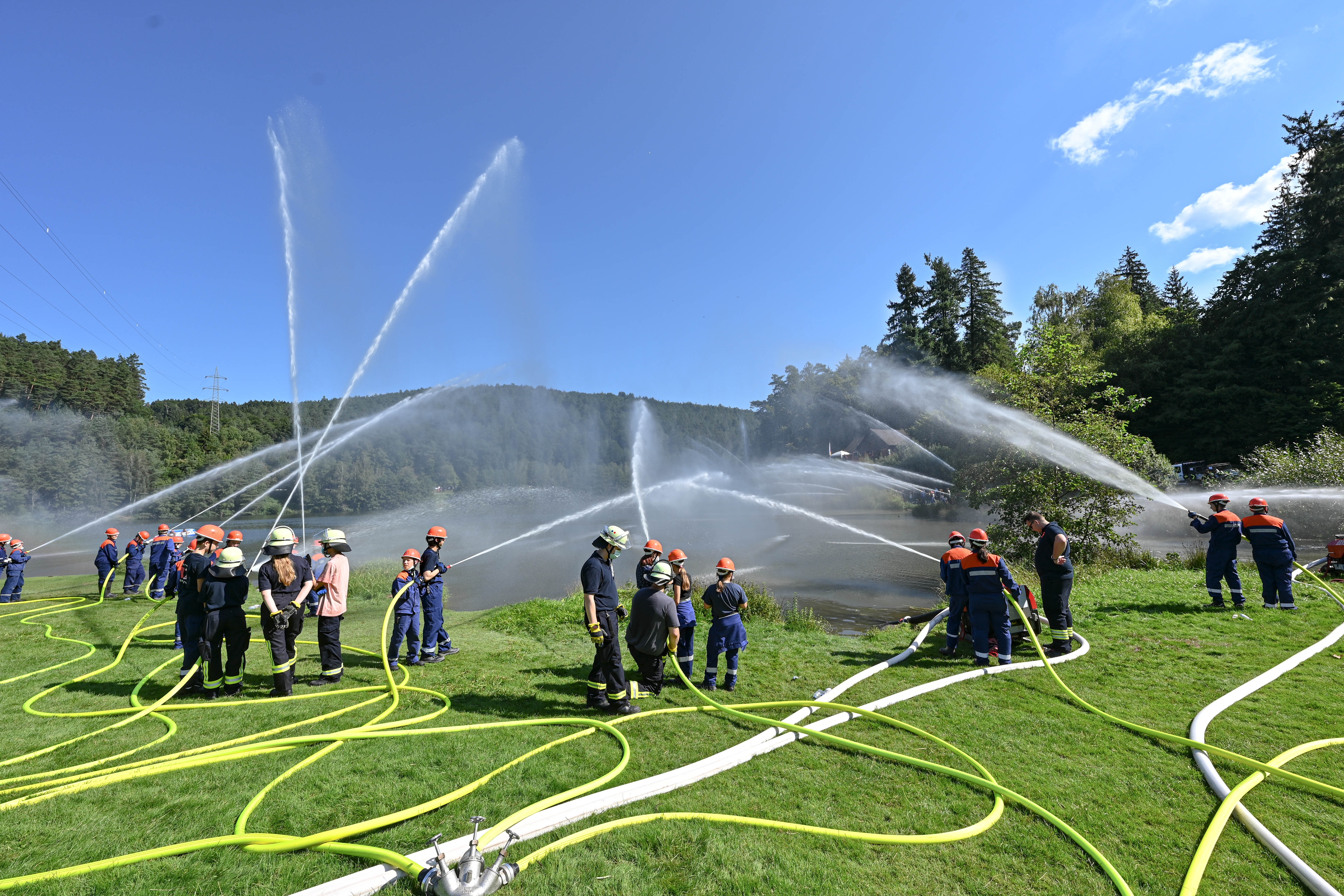 Das Bild zeigt Kinder der Feuerwehr, die mit Schläuchen Wasser in einen See spritzen.
